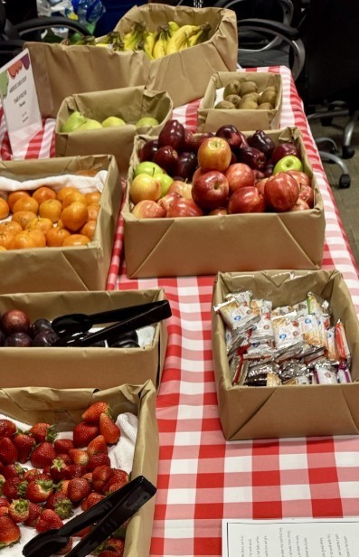  Table display of fresh fruit and packaged snacks on a red-and-white checkered tablecloth. Cardboard trays hold bananas, green pears, kiwis, clementines/tangerines, mixed apples and plums, dark purple plums (with tongs), and strawberries (with tongs). Individually wrapped snack bars sit among the fruit. A sign nearby reads "APPLE-A-DAY HEALTHY SNACK PROGRAM" and a poster explains fruit is nutritious, hydrating, and gives energy to tackle the day. The scene conveys a healthy snack station set up indoors.