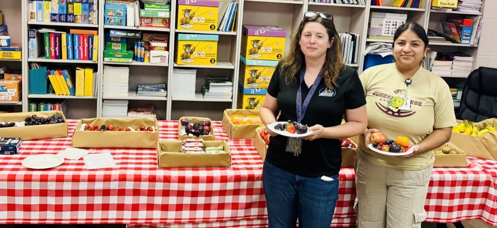 Two women stand behind a table covered with a red-and-white checkered tablecloth holding plates of fresh fruit. The table displays trays of grapes, strawberries, plums, oranges/tangerines, apples, and bananas, plus some packaged snack bars. Behind them, white bookshelves hold binders, boxed learning kits (including "Fundations Wilson Language Program"), board games, and labeled materials. Both women smile at the camera; one wears a black shirt and sunglasses on her head, the other wears a tan "EUREKA COLLEGE" shirt. The scene shows a community group providing a fresh food spread in a classroom or resource room.
