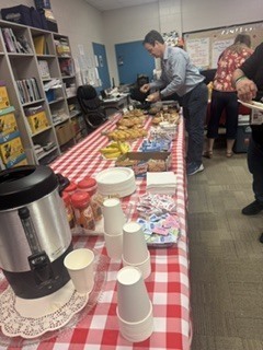 Indoor buffet scene with a long table covered in a red-and-white checkered tablecloth holding pastries, muffins, individually wrapped snacks, a large silver coffee dispenser, stacks of disposable white cups, and small red condiment containers. A man in a light blue shirt is serving himself while others gather nearby; shelving with binders and office supplies is visible to the left. The image conveys a communal breakfast or staff event.