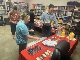 Adults gathered around tables covered with red-and-white checkered tablecloths serving themselves a casual buffet of hot dogs or sausages, buns, and condiments. One person places items into containers while another serves from a platter. Shelving with books and binders is visible in the background, and a banner reads "WE ♥ OUR TEACHERS & STAFF!" The scene appears to be a staff appreciation meal in an office or breakroom.