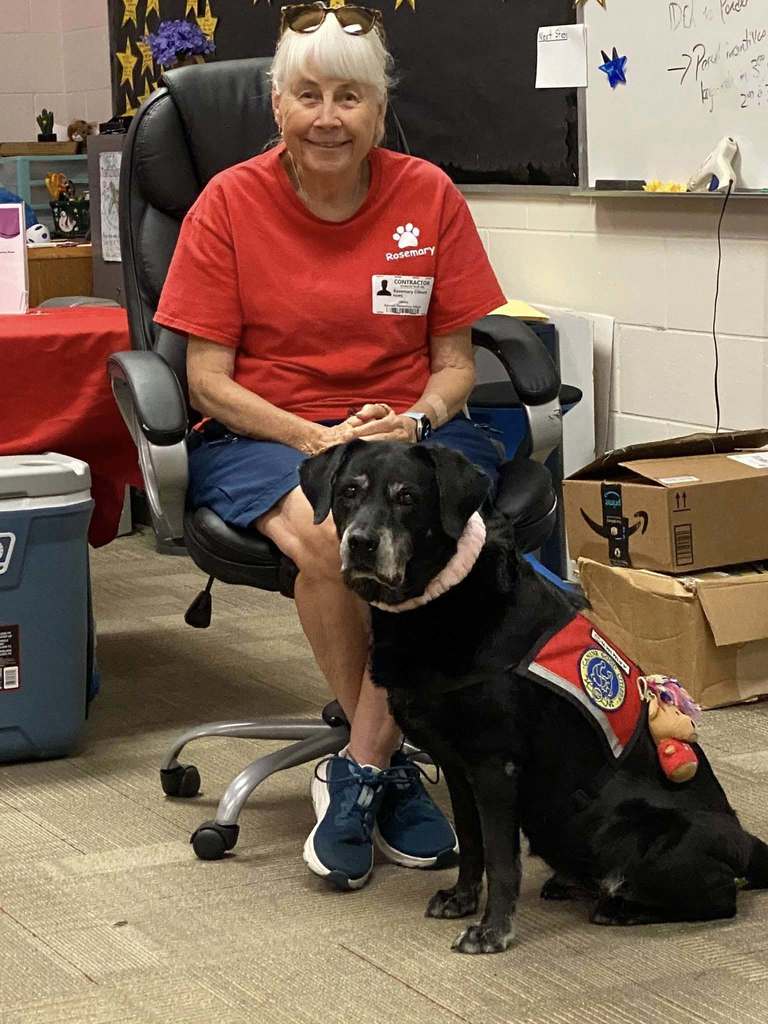 Photo of a woman in a red shirt who is holding the collar of a black medium sized dog. 