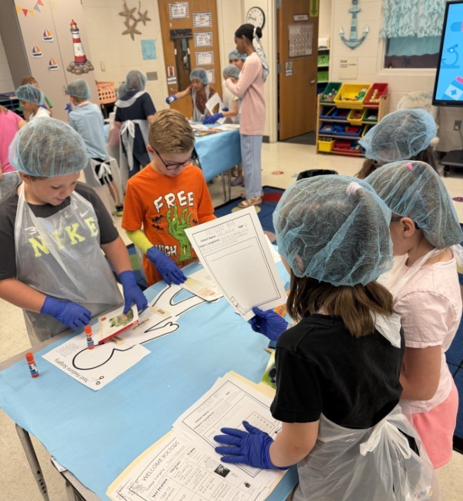 Elementary students wearing light-blue disposable gowns, hairnets, and shoe covers work at white tables inside makeshift cubicles formed by light-blue drapes. Red-and-white "RESTRICTED AREA AUTHORIZED EMPLOYEES -ONLY-" signs are posted on the drapes. Classroom details—storage cabinets, a large flat-screen TV, educational posters including numbers 1–10 and a "MATH" poster, a wall clock, and a dark gray shag rug in the aisle—are visible in the background. The scene resembles a simulated lab or role-play activity.
