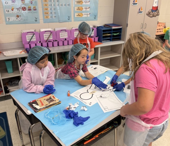Elementary students wearing light-blue disposable gowns, hairnets, and shoe covers work at white tables inside makeshift cubicles formed by light-blue drapes. Red-and-white "RESTRICTED AREA AUTHORIZED EMPLOYEES -ONLY-" signs are posted on the drapes. Classroom details—storage cabinets, a large flat-screen TV, educational posters including numbers 1–10 and a "MATH" poster, a wall clock, and a dark gray shag rug in the aisle—are visible in the background. The scene resembles a simulated lab or role-play activity.