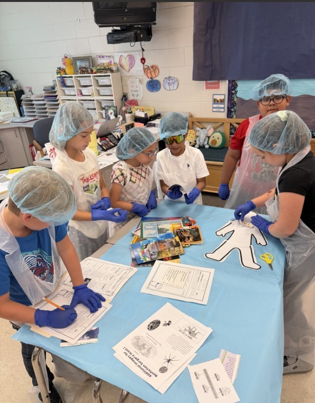 Elementary students wearing light-blue disposable gowns, hairnets, and shoe covers work at white tables inside makeshift cubicles formed by light-blue drapes. Red-and-white "RESTRICTED AREA AUTHORIZED EMPLOYEES -ONLY-" signs are posted on the drapes. Classroom details—storage cabinets, a large flat-screen TV, educational posters including numbers 1–10 and a "MATH" poster, a wall clock, and a dark gray shag rug in the aisle—are visible in the background. The scene resembles a simulated lab or role-play activity.