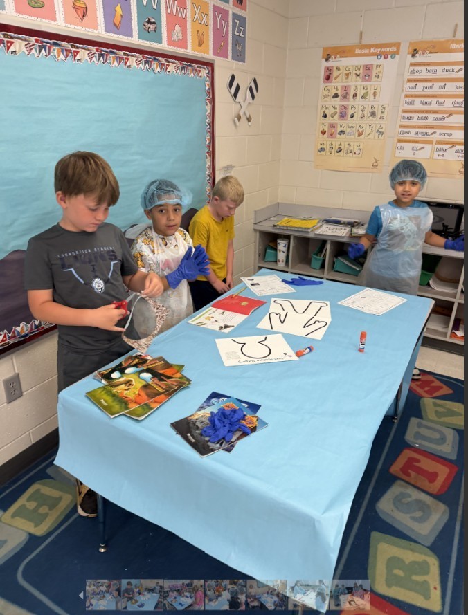 Elementary students wearing light-blue disposable gowns, hairnets, and shoe covers work at white tables inside makeshift cubicles formed by light-blue drapes. Red-and-white "RESTRICTED AREA AUTHORIZED EMPLOYEES -ONLY-" signs are posted on the drapes. Classroom details—storage cabinets, a large flat-screen TV, educational posters including numbers 1–10 and a "MATH" poster, a wall clock, and a dark gray shag rug in the aisle—are visible in the background. The scene resembles a simulated lab or role-play activity.