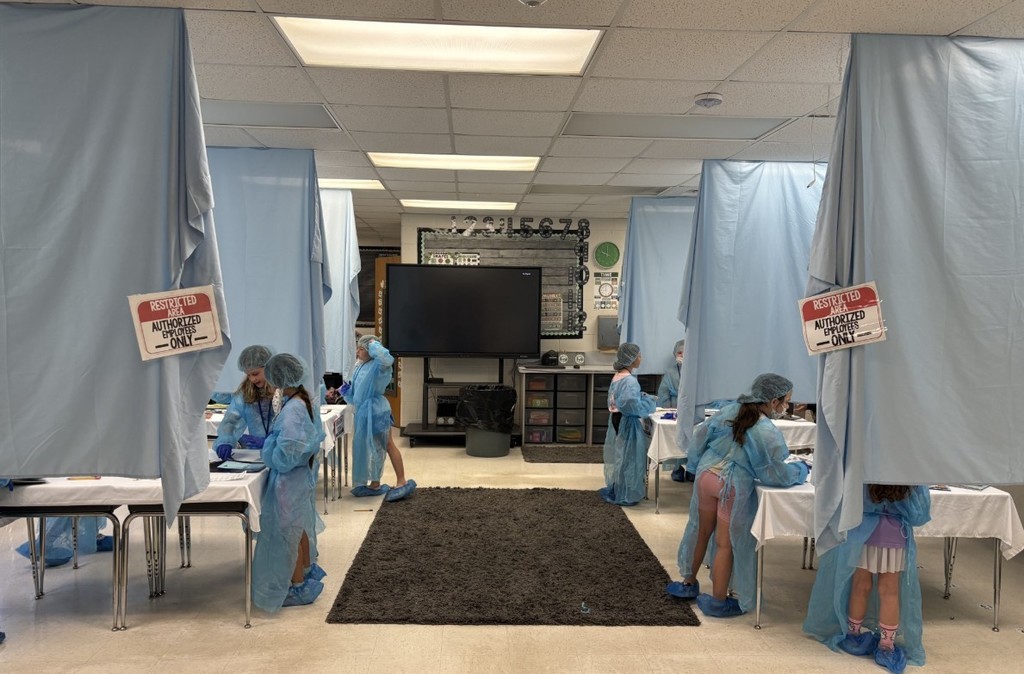 Elementary students wearing light-blue disposable gowns, hairnets, and shoe covers work at white tables inside makeshift cubicles formed by light-blue drapes. Red-and-white "RESTRICTED AREA AUTHORIZED EMPLOYEES -ONLY-" signs are posted on the drapes. Classroom details—storage cabinets, a large flat-screen TV, educational posters including numbers 1–10 and a "MATH" poster, a wall clock, and a dark gray shag rug in the aisle—are visible in the background. The scene resembles a simulated lab or role-play activity.