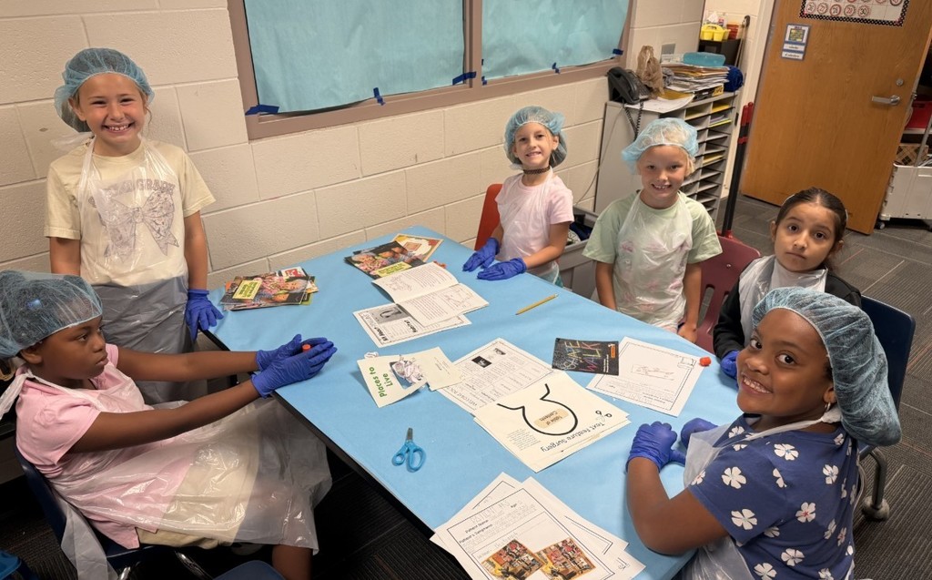 Elementary students wearing light-blue disposable gowns, hairnets, and shoe covers work at white tables inside makeshift cubicles formed by light-blue drapes. Red-and-white "RESTRICTED AREA AUTHORIZED EMPLOYEES -ONLY-" signs are posted on the drapes. Classroom details—storage cabinets, a large flat-screen TV, educational posters including numbers 1–10 and a "MATH" poster, a wall clock, and a dark gray shag rug in the aisle—are visible in the background. The scene resembles a simulated lab or role-play activity.