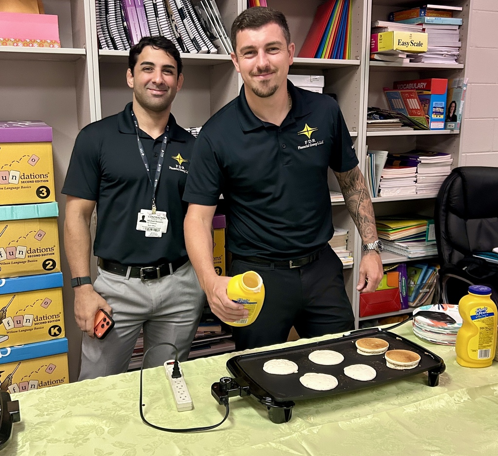 Two men stand behind a table with an electric griddle, making pancakes in a classroom or office setting. The man on the left wears an ID badge and holds a smartphone; the man on the right, with visible arm tattoos, holds a yellow Shake ’n Pour buttermilk pancake mix bottle and appears to pour batter onto the griddle where several pancakes are cooking. The table has a light green tablecloth, a power strip, and cooking supplies. Behind them are bookshelves filled with educational materials, including stacked "Fundations — Wilson Language Basics (Second Edition)" boxes labeled K, 2, and 3 on the left, and books/binders such as "Easy Scale" and "VOCABULARY" on the right.