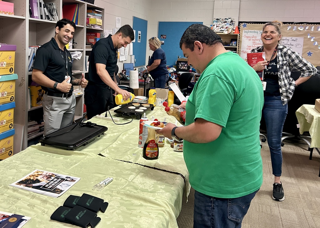 Indoor pancake breakfast scene with five adults gathered around a long table covered in a light green tablecloth. Two men in black polos are near the griddles — one smiling and giving a thumbs-up, the other pouring pancake batter — while a man in a green t-shirt at the table end adds toppings (syrup, whipped cream, strawberries) to a plated pancake. In the background, a woman in a plaid shirt laughs holding a red folder and another woman in blue is partially visible. The table holds two electric griddles, pancake ingredients, syrup (Pearl Milling Company), a jar of Nutella, promotional koozies, and flyers. Shelves of instructional materials (including "Foundations / Language Basics" boxes for grades K, 2, 3) and a bulletin board with notes are visible behind them.
