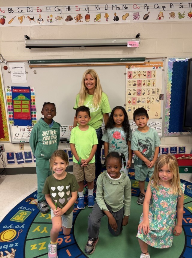 Photo of a classroom group portrait: a smiling female teacher stands at the back wearing a light green T‑shirt and jeans; seven young children sit and kneel in front of her, most dressed in green or green‑toned clothing. The classroom walls behind them display colorful educational charts and posters, including an alphabet strip with pictures, a large word‑family chart (examples: -old, -all, -ake, -ick, -ing, -ank, -ed, -est, -eat, -in, -ip, -it), a phonics keywords chart (Aa apple through Zz zebra and lists of digraphs, vowel teams, r‑controlled vowels, and ending sounds), and a partial number chart. A circular rug with letters and numbers is on the floor. The scene conveys a cheerful elementary classroom participating in a “Be Seen in Green” activity for Mental Health Awareness Month.