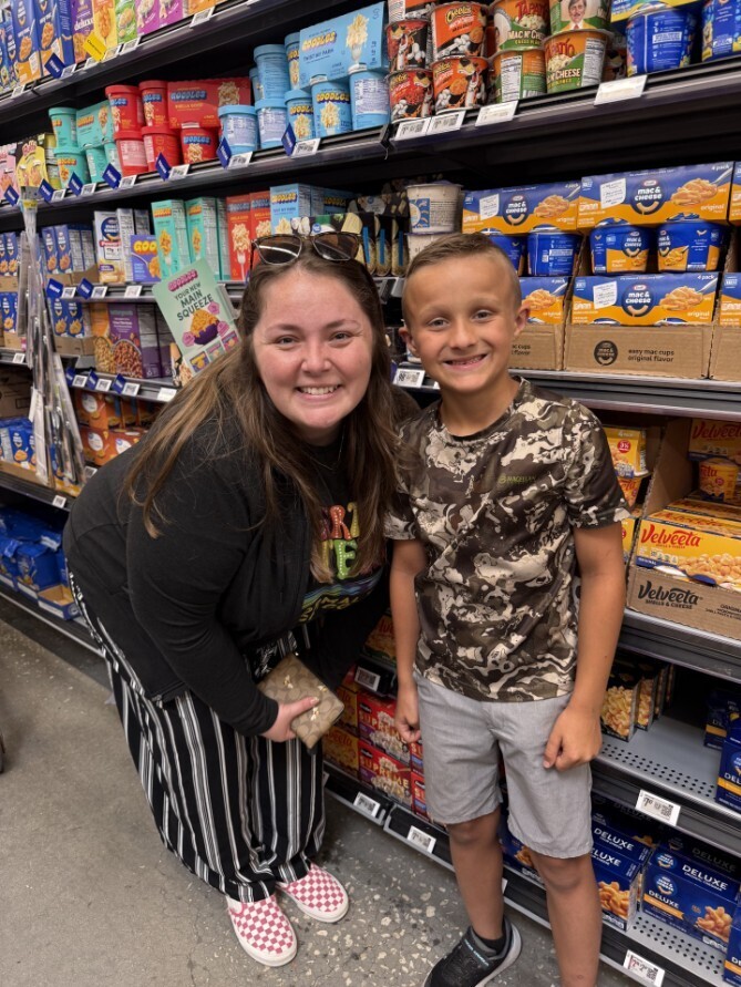 A photo taken in a grocery store aisle showing a smiling woman and young boy standing side by side. Behind them, shelves are stocked with boxed and cup mac & cheese, popcorn tubs, and snack cups. The woman has long brown hair, wears a black long-sleeve top over a colorful shirt, black-and-white striped pants, and pink-and-white checkered slip-on shoes; she holds a small patterned wallet. The boy has short blond hair, a camouflage T‑shirt with a partially visible “MAGELLAN” label, gray shorts, and black sneakers. Visible product labels include Doodles Sweet ’N Salty Caramel Popcorn, Cheetos Mac ’N Cheese cups, Velveeta Mac & Cheese (cups and boxes), Kraft Mac & Cheese and other instant mac & cheese varieties. The woman holds a pink display card reading “Goo” and “Your New Main Squeeze.” The scene is well lit and focused on the people and nearby grocery products.