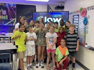 Group photo in a classroom with dim blue lighting and a large screen behind reading “Glow.” About 15 children and one adult pose together; many wear neon or brightly colored shirts and glow accessories like bracelets. Several children and the adult give thumbs up, conveying excitement and celebration of the themed event.