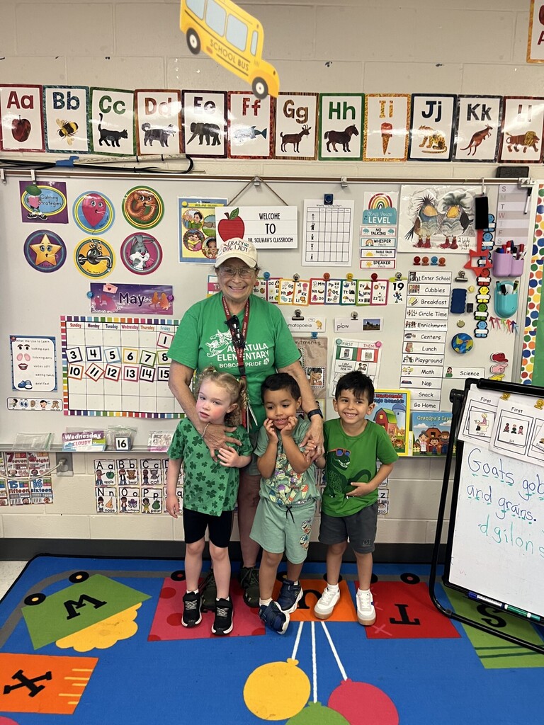  Vibrant elementary classroom with colorful alphabet cards and learning posters on a whiteboard. A smiling teacher wearing a green “AATULA ELEMENTARY BOBCA PRIDE” T‑shirt and a white “CRAZY DOG LADY” cap stands with three young children, all wearing green shirts. The whiteboard displays a May calendar, a “What We Use To Learn” poster (looking eyes, listening ears, voices soft, waiting hands, kind feet), a voice level chart, and a daily schedule. A blue rug with letters and bright shapes covers the floor; a yellow school bus cutout hangs from the ceiling.