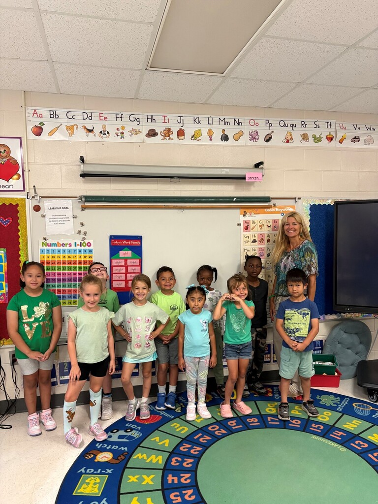 Classroom photo of a female teacher standing with nine young students on a colorful circular alphabet rug. The teacher wears a patterned blouse; the diverse group of children stand in front of her smiling at the camera. Alphabet strips and educational charts line the walls. A whiteboard shows a learning goal about phonemic awareness and a numbers 1–100 chart; bright, welcoming classroom environment.