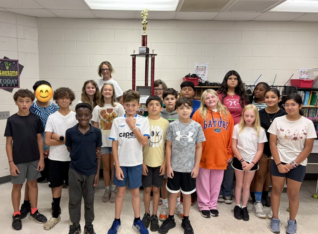 A group portrait of about 20 diverse preteen children standing in rows indoors with an adult not visible. A large two-tiered trophy with red columns and a gold figure tops a dark base and sits behind the group. The room has a light brick-patterned wall, a green poster on the left reading “Be Awesome TODAY,” and shelves with books and school supplies on the right labeled “LEFT SIDE” and “RIGHT SIDE.” A partially obscured poster behind the trophy reads “COMPETING CLASS OF THE WEE[K] GREAT.” Children wear casual clothes with visible text and logos (examples: a white jersey reading “Capelli cape SPORTS,” an orange hoodie reading “FLORIDA GATORS,” a dark pink t‑shirt reading “NEW YORK CITY AEROPOSTALE,” a gray camouflage Under Armour shirt, and a yellow t‑shirt with “OX” in the design).