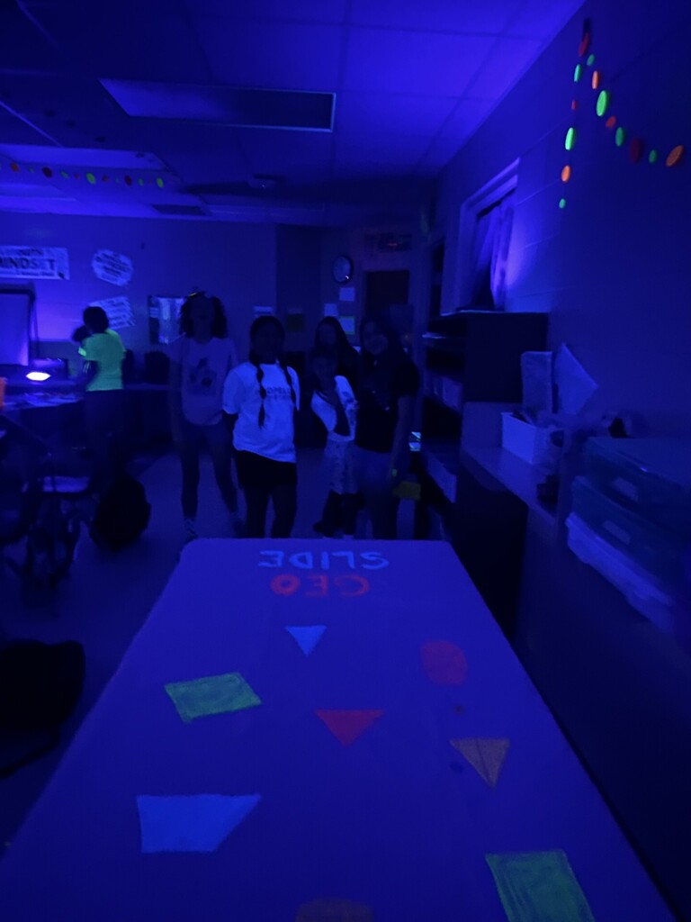 A classroom bathed in blue/purple blacklight with a disco-ball–style light casting red, green, and blue patterns on the ceiling. Neon paper garlands in bright pink, green, and orange hang across the drop ceiling and glow vividly. A bright white light source and various ceiling vents and fixtures are visible. A wall clock and directional signs appear on the walls; one wall shows the quote “ALL THINGS ARE DIFFICULT before they are EASY.” The overall scene shows decorations and lighting for a glow event.