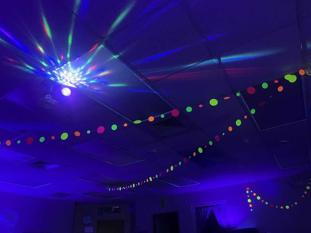 A blacklight-lit room with students and bright neon decorations. Walls are trimmed with glowing circular and oval shapes in green, orange, red, and yellow. In the foreground, a table displays glowing geometric cutouts (a green parallelogram, blue trapezoid, red/light-blue/yellow triangles, and a red circle) and a sign reading “GEO SLIDE” (upside down from viewer). One person on the left wears a neon yellow shirt that fluoresces under the UV light. A wall sign partly reads “MINDSET.” The scene suggests a glow-themed, geometry-focused learning station.