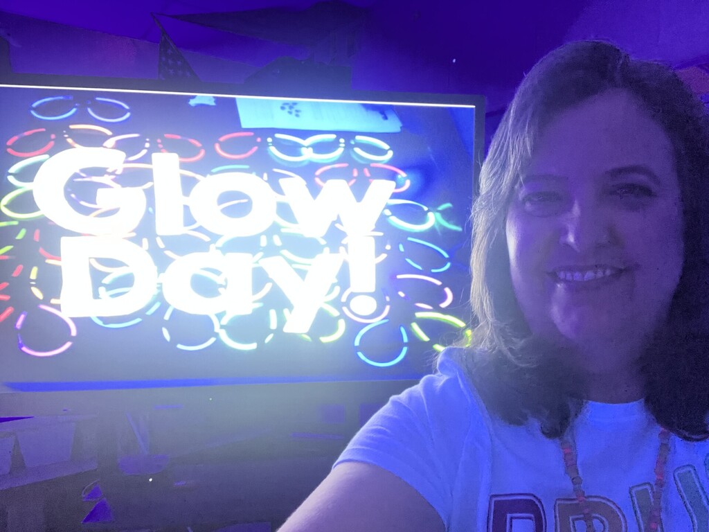 A person with long dark hair smiles for a selfie in a dim room lit by purple/blue blacklight. They wear a white T-shirt and a beaded necklace with some glowing beads. To the left, a monitor or TV screen displays the words “Glow Day!” in bright white letters surrounded by many colorful glowing circles (blue, red, green, yellow, pink, orange), suggesting glow sticks or rings. The scene feels festive and glow-themed.