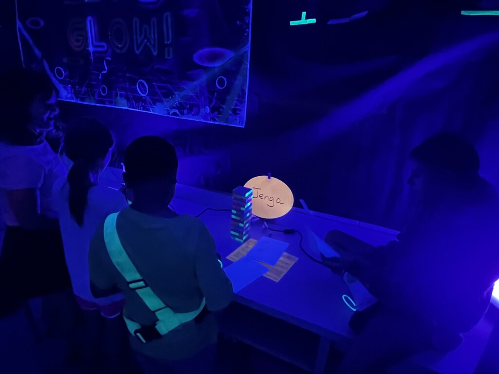  A group of children or teens in a dim blacklight room where neon blues and greens glow. In the center, a person seen from behind wears a glowing green strap. On a table in front, a stack of Jenga blocks glows; a circular sign above the table reads “Jenga.” The back wall features glowing patterns and the word “GLOW!” Ceiling T-shaped glowing elements and glowing cards or papers on the table suggest a glow-themed game night.