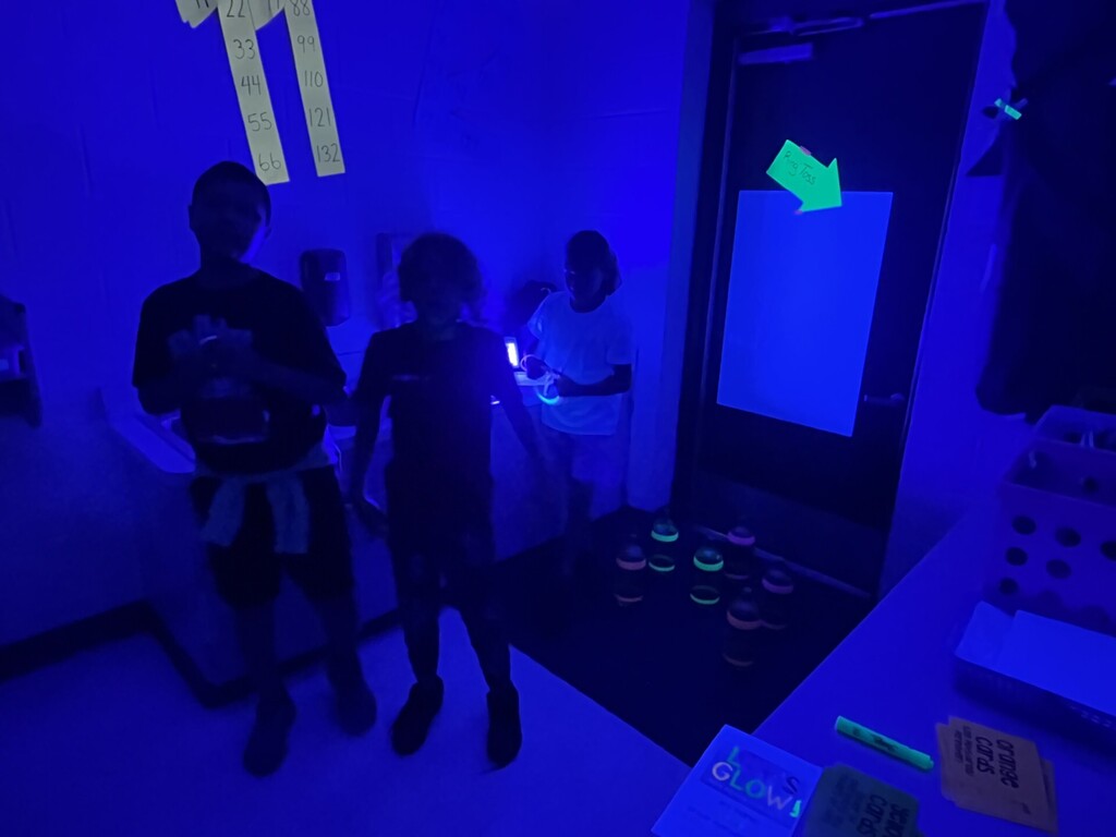  A room lit by blue UV light showing activity stations and game materials. Paper strips with numbers (11, 22, 33, 44, 55, 66, 88, 99, 110, 121, 132) are taped to a wall. A glowing green arrow on a door points down to “Ring Toss.” A box or sign in the foreground reads “IT’S GLOW!” Stacks of labeled cards (“ORANGE CARDS,” “SPUD CARDS,” and partially visible green cards) and glow sticks sit on a table. Three people stand in the room; several dark bottles with glowing green tops are arranged on the floor, likely for a game. The image conveys a glow-in-the-dark event with organized stations.