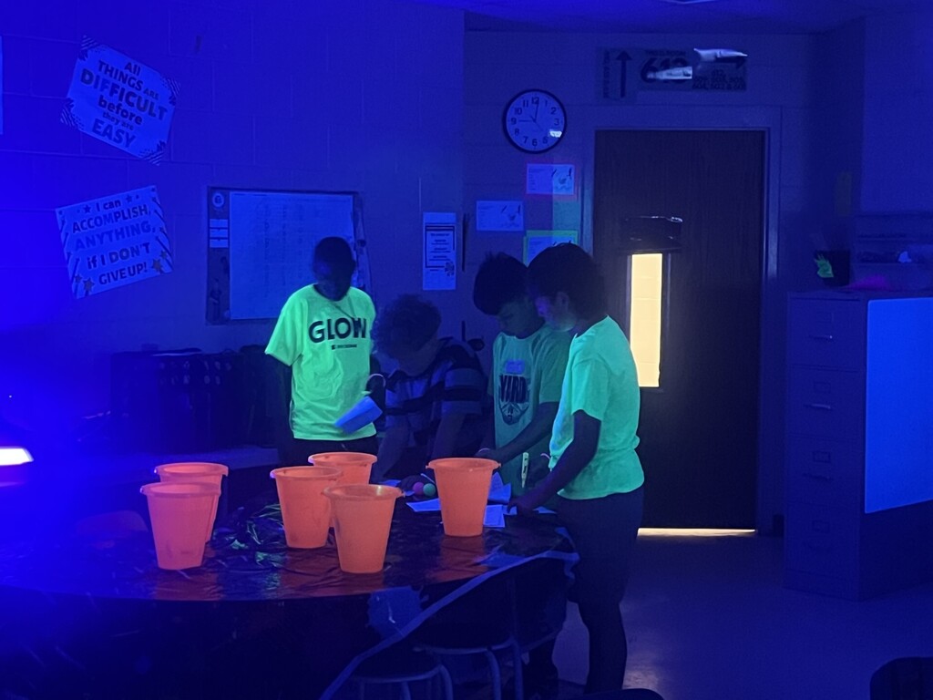 Four children gather around a table in a classroom lit by blacklight, creating a strong blue hue. The table holds glowing orange cups and small glowing balls in green and pink/orange. Three boys wear fluorescent neon shirts (one shirt reads “GLOW”), which fluoresce brightly under the UV light. A wall clock (about 12:08–12:10), motivational posters, a dark door with a lit window, and a filing cabinet are visible in the background. A sign above a doorway displays motivational text: “ALL THINGS ARE DIFFICULT before they are EASY” and a classroom sign reading “THIS ROOM 610 MRS. / MR. [line]’S CLASS.”