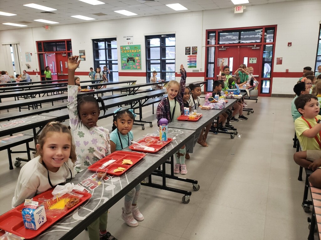 Elementary school cafeteria during lunchtime showing a diverse group of students seated at long grey tables with red lunch trays; many children are smiling and waving at the camera. Foreground details include a boy in a green-and-white striped shirt raising his hand and a girl in a tie-dye shirt beside a Hello Kitty lunchbox. Visible signage includes a large red circular school logo with a wolf/dog mascot, EXIT signs, clocks, and milk cartons on trays. Bright natural and overhead lighting and supervising adults create a safe, active learning environment.