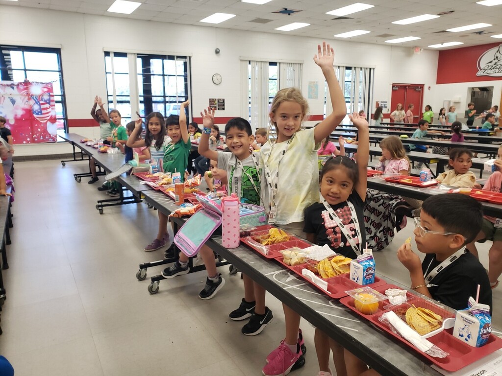 Elementary school cafeteria during lunchtime showing a diverse group of students seated at long grey tables with red lunch trays; many children are smiling and waving at the camera. Foreground details include a boy in a green-and-white striped shirt raising his hand and a girl in a tie-dye shirt beside a Hello Kitty lunchbox. Visible signage includes a large red circular school logo with a wolf/dog mascot, EXIT signs, clocks, and milk cartons on trays. Bright natural and overhead lighting and supervising adults create a safe, active learning environment.