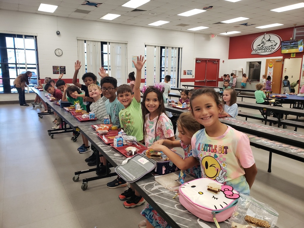 Elementary school cafeteria during lunchtime showing a diverse group of students seated at long grey tables with red lunch trays; many children are smiling and waving at the camera. Foreground details include a boy in a green-and-white striped shirt raising his hand and a girl in a tie-dye shirt beside a Hello Kitty lunchbox. Visible signage includes a large red circular school logo with a wolf/dog mascot, EXIT signs, clocks, and milk cartons on trays. Bright natural and overhead lighting and supervising adults create a safe, active learning environment.