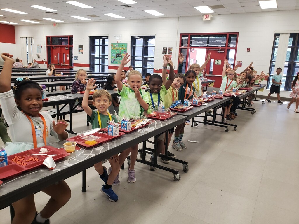 Elementary school cafeteria during lunchtime showing a diverse group of students seated at long grey tables with red lunch trays; many children are smiling and waving at the camera. Foreground details include a boy in a green-and-white striped shirt raising his hand and a girl in a tie-dye shirt beside a Hello Kitty lunchbox. Visible signage includes a large red circular school logo with a wolf/dog mascot, EXIT signs, clocks, and milk cartons on trays. Bright natural and overhead lighting and supervising adults create a safe, active learning environment.