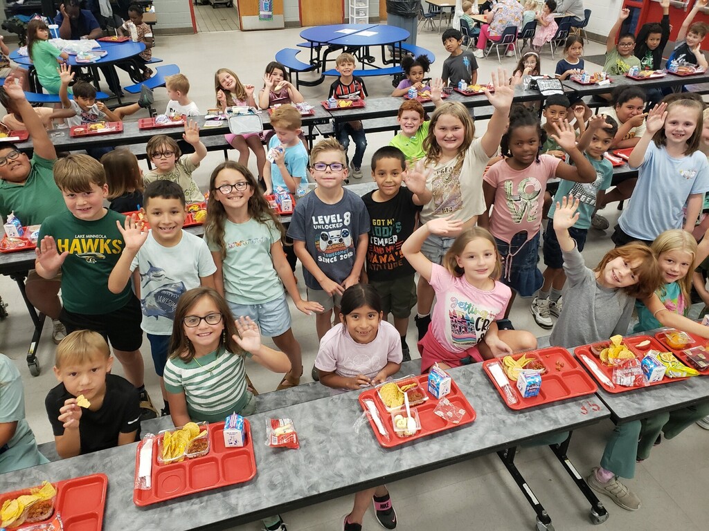 Elementary school cafeteria during lunchtime showing a diverse group of students seated at long grey tables with red lunch trays; many children are smiling and waving at the camera. Foreground details include a boy in a green-and-white striped shirt raising his hand and a girl in a tie-dye shirt beside a Hello Kitty lunchbox. Visible signage includes a large red circular school logo with a wolf/dog mascot, EXIT signs, clocks, and milk cartons on trays. Bright natural and overhead lighting and supervising adults create a safe, active learning environment.