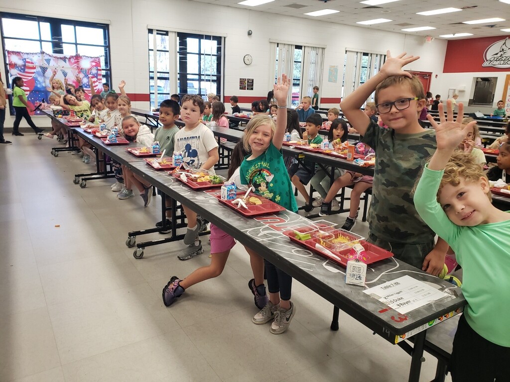 Elementary school cafeteria during lunchtime showing a diverse group of students seated at long grey tables with red lunch trays; many children are smiling and waving at the camera. Foreground details include a boy in a green-and-white striped shirt raising his hand and a girl in a tie-dye shirt beside a Hello Kitty lunchbox. Visible signage includes a large red circular school logo with a wolf/dog mascot, EXIT signs, clocks, and milk cartons on trays. Bright natural and overhead lighting and supervising adults create a safe, active learning environment.