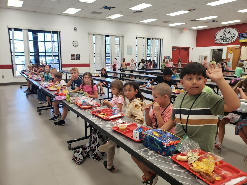 Elementary school cafeteria during lunchtime showing a diverse group of students seated at long grey tables with red lunch trays; many children are smiling and waving at the camera. Foreground details include a boy in a green-and-white striped shirt raising his hand and a girl in a tie-dye shirt beside a Hello Kitty lunchbox. Visible signage includes a large red circular school logo with a wolf/dog mascot, EXIT signs, clocks, and milk cartons on trays. Bright natural and overhead lighting and supervising adults create a safe, active learning environment.
