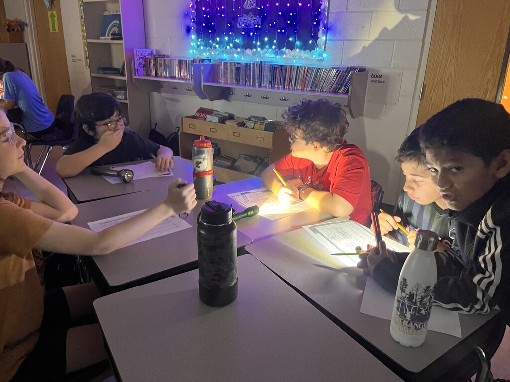 Five students gathered around two tables in a dimly lit classroom, each using flashlights to read and write on papers. Some students hold flashlights close to worksheets; others write by the beam. Background shows bookshelves, classroom storage, and a board with blue-and-white string lights and a "WE LEARN" sign. Visible text on objects includes "VESTIBULE," "60BA," and brand labels like "Gatorade" and "STRANGER THINGS" on water bottles. A person in a blue shirt is partially visible at the far left.