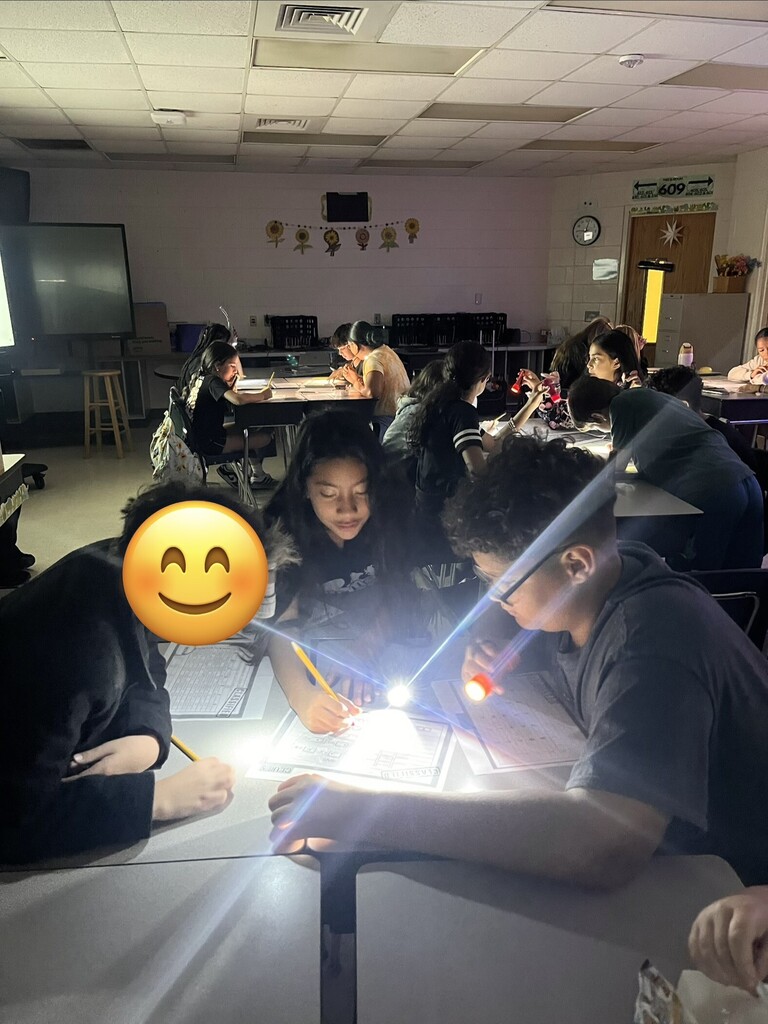 Dimly lit classroom where students sit at individual desks using flashlights to illuminate worksheets. Ceiling acoustic tiles and vents are visible; a large projection screen or whiteboard sits at left. Back wall features a row of five sunflower cutouts, a clock, and a door with posted emergency contact text above it. Several students hold flashlights; one foreground student uses a red flashlight at right. A yellow smiling emoji overlays a student’s face in the lower-left foreground. Visible wall text above the door lists emergency numbers (POLICE 911, AMBULANCE 911, FIRE 911) and other unreadable phone numbers. A partially visible worksheet labeled with a form-like title (e.g., "EXAMINER II") appears on a desk.