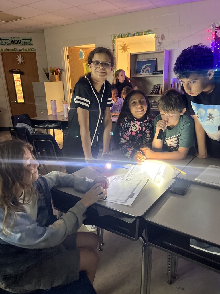 Group of elementary/middle school students working in a dim classroom using flashlights to read papers. Foreground center: a girl in a tie-dye long-sleeve shirt holds a red flashlight over a paper labeled "CODE TO DE-BUG." Nearby, a girl with glasses and curly hair in a black-and-white baseball jersey smiles at the camera, while other students sit or lean over desks holding flashlights. Wall text above a door reads "THIS IS ROOM 609" with directions or room numbers beneath ("GO 412, 510, 615, & 616"). Classroom features include bookshelves, a framed rainbow picture, purple string lights, student desks, and a star on a door.