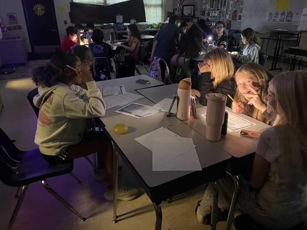 Classroom scene in low light with small groups of students working at gray desks pushed together. Most students use phone flashlights or small lamps to read papers on their desks. Foreground left: a student with dark curly hair in a gray hoodie (partial "AKAN" text) leans over papers with a yellow spherical object beside them. Foreground right: two blonde students at a desk — one in a black top looking down at illuminated papers, the other smiling and holding a pencil; a pink "owala" water bottle, light pink tumbler, and phone sit nearby. Mid-ground and background: more students focused on worksheets by flashlight; one student on the right looks toward the camera brightly lit. Classroom details include light-colored walls, a sunflower decoration on a door, an American flag, and windows with dark blinds. Papers with grids and text are visible but unreadable from this distance.