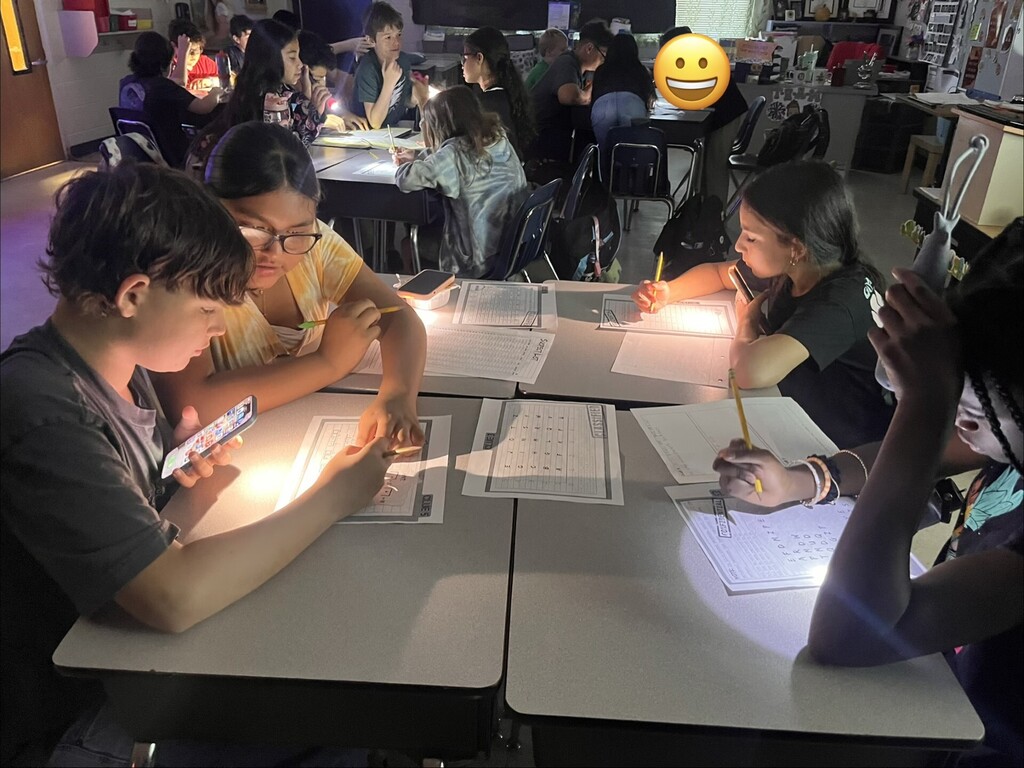 Low-light classroom activity with students using phone flashlights and small lamps to work on worksheets. Foreground: a boy shines his phone flashlight at his work beside a girl wearing glasses; both focus on separate papers. To their right, girls use individual light sources — one with a flexible desk lamp held over her worksheet. Background shows more students similarly engaged. A yellow smiling emoji covers a distant student’s face. Visible worksheet text includes headings like "CLUES" with numbered lines, a "SUPPLY" grid containing letters (e.g., TISSUE) and numbers, and "TRANSFORM" grids with letter sequences; grid details are partially visible but not fully readable.