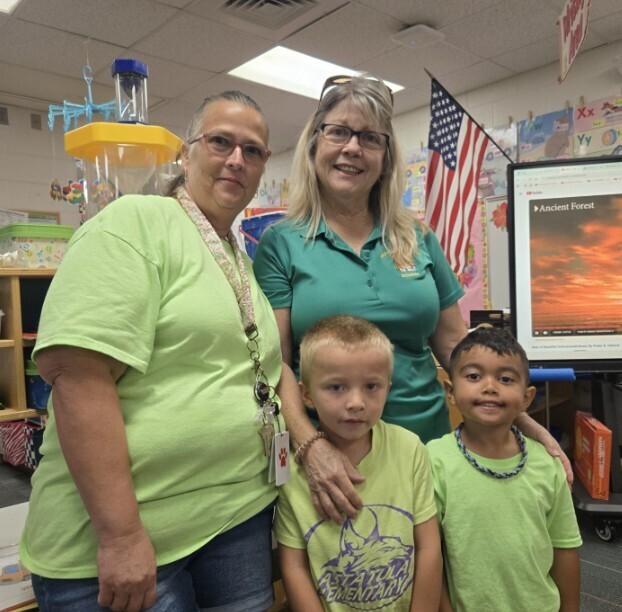 Four people—two adult women and two young boys—pose in a classroom, all wearing green shirts. The woman on the left wears a light green t-shirt, denim shorts, glasses, and a lanyard; the woman on the right wears a dark green polo, glasses, and sunglasses on her head, with her hand on the shoulder of the boy beside her. The two boys stand in front: the left boy wears a light green shirt printed with "ASTATOULA ELEMENTARY," and the right boy wears a light green shirt and a dark beaded necklace. Behind them an American flag and a large screen display a YouTube video titled "Ancient Forest" showing a scenic forest sunset; classroom shelves and decorations are visible.