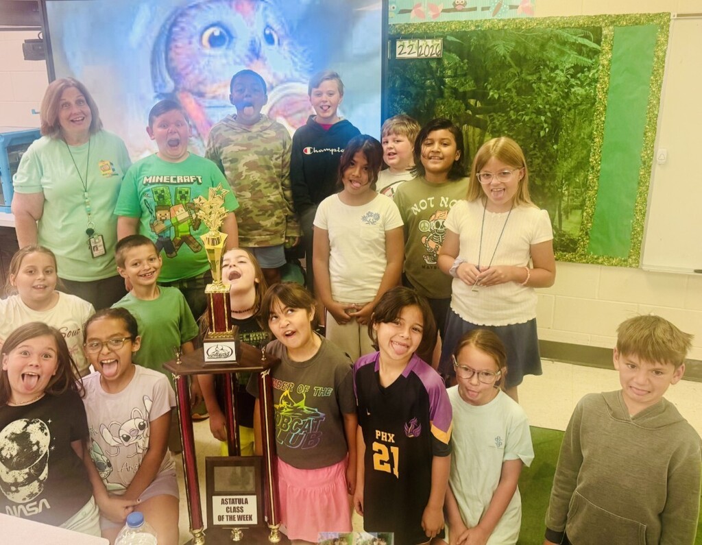 Group photo in a classroom showing one adult woman (teacher) and a diverse group of smiling elementary students gathered around a large gold trophy. The trophy base plaque reads "ASTATULA CLASS OF THE WEEK." Children wear a variety of graphic shirts (examples visible: MINECRAFT, Champion, NOT NO MAYBE with a skeleton graphic, NASA, Angel with a Stitch character, PHX 21, and LEADER OF THE BOBCAT CLUB). Background includes a decorative display with an owl image and a dark green forest motif; the numbers "222020" appear on the display. The mood is celebratory and proud.