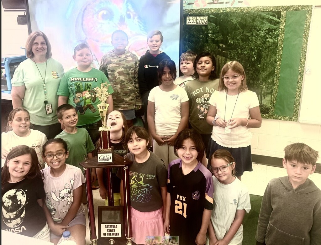 Group photo in a classroom showing one adult woman (teacher) and a diverse group of smiling elementary students gathered around a large gold trophy. The trophy base plaque reads "ASTATULA CLASS OF THE WEEK." Children wear a variety of graphic shirts (examples visible: MINECRAFT, Champion, NOT NO MAYBE with a skeleton graphic, NASA, Angel with a Stitch character, PHX 21, and LEADER OF THE BOBCAT CLUB). Background includes a decorative display with an owl image and a dark green forest motif; the numbers "222020" appear on the display. The mood is celebratory and proud.