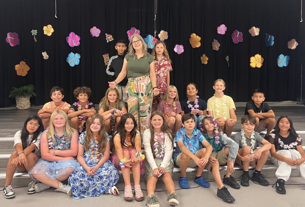 Group photo of one adult woman and many children posing on tiered steps in a school setting. The backdrop is a black curtain decorated with colorful hibiscus flower cutouts and small palm trees, giving a luau or tropical theme. The adult (wearing glasses, an olive-green top, and patterned pants) stands near center-left; most children sit on the steps in front, many wearing leis and smiling at the camera. The scene conveys a celebratory, joyful moment honoring longtime Bobcats who began in Pre‑K with Mrs. Quero.