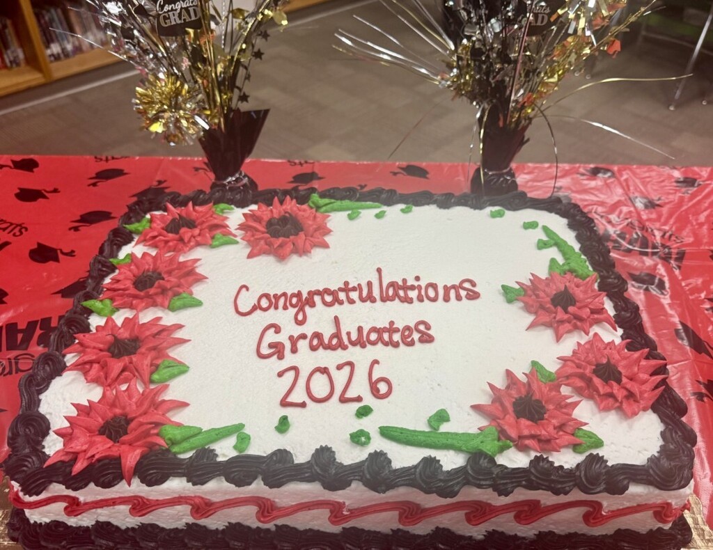 Rectangular white frosted celebration cake decorated with red icing flowers and green leaves, a black piped top border, and a red-and-white wavy base border. Red icing on top reads "Congratulations Graduates 2026." The cake sits on a red tablecloth patterned with black graduation caps; behind it are two festive centerpieces with metallic gold and silver tinsel and small "CONGRATS GRAD" cutouts.