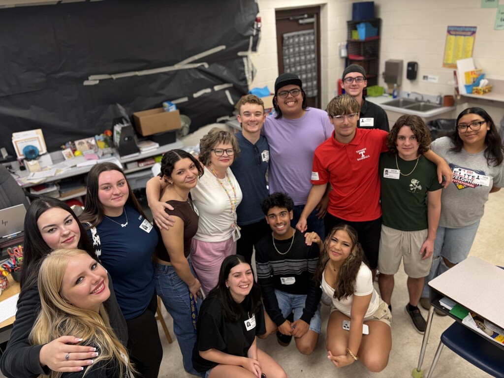Classroom photo of 12 students seated at individual desks with one adult teacher standing behind them. Students mostly smile at the camera; several desks show name tags (examples: NOVA, SARA, SadieR, Landon, Skyler, Jude, L. EMMANUEL, Mason). Classroom walls display numeric ranges and posters; a clock, filing cabinet, and two wooden doors are visible on the right. A front-right desk holds a small plaque reading "Pamela Duke IT Teacher of the Year 2013–2014" and other miscellaneous items. The image captures a bright, organized learning environment and a group school photo.