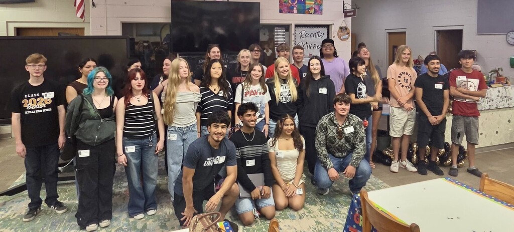 Large group photo of diverse high school seniors gathered indoors, most standing with a few crouching in front. Many wear ID badges. Decorations include gold and black balloons, trophies on the floor at right, and framed artwork on the walls. An American flag is visible at upper left. A prominent sign reads "CLASS OF 2026 SENIOR" and references "AYTILLA HIGH SCHOOL" and "Recently Returned Welcome," indicating this is a reunion or reception for returning alumni.