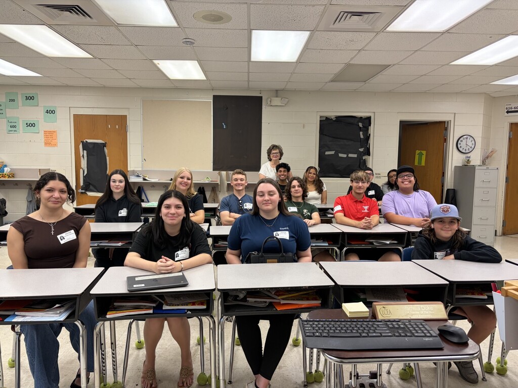 Group photo of 16 mostly young adults and teens with one older woman, posed and smiling in a classroom/office setting. Left foreground shows a cluttered desk with an open laptop (HP logo), papers, and supplies; the wall above is covered in black material. Right background includes a sink, countertops, storage bins, and a closed door. Visible clothing text includes "SAVAGER BASEBALL" on a red polo, "JOSTEN" on a name tag, and "USA ELITE! VELOPME INVITATIONAL" on a gray T‑shirt. The scene feels casual and celebratory, likely documenting a school gathering or alumni return.
