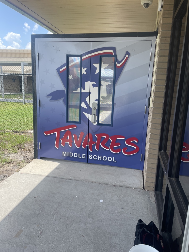 Double doors with a large blue sign reading "TAVARES MIDDLE SCHOOL" are open. A security camera is visible at the top of the image.