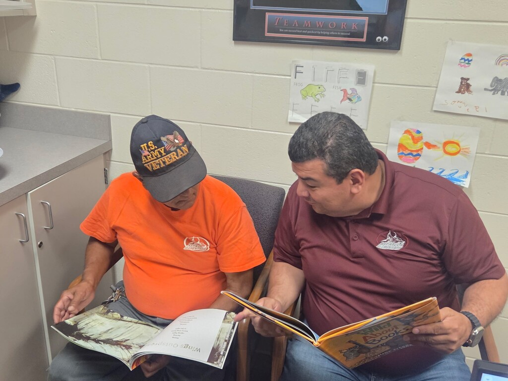 Mr. Carlos and Mr. Shorty are sitting in chairs and reading children's books in the office.