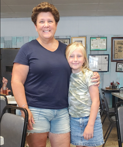 Woman and girl stand close. The woman wears a blue shirt and denim shorts. The girl has short hair and wears a camo shirt.