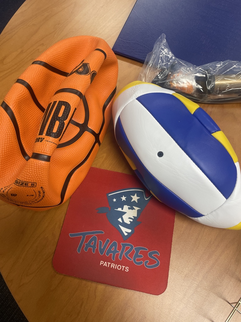 Items on a table: an orange basketball glove, a blue and white ball, a red coaster with "TAVARES PATRIOTS" text, and a blue plastic sheet.