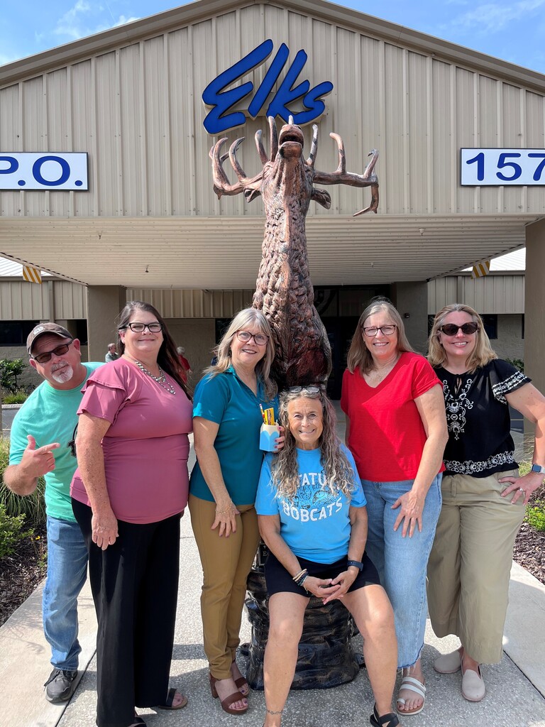Six adults pose for a photo in front of a building with an Elk statue.